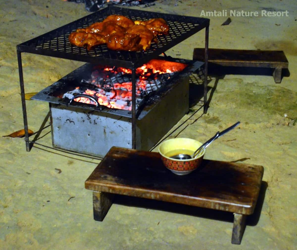 Charcoal barbecue at night with marinated chicken pieces on the upper grill rack over glowing coals, a low wooden stool alongside holding a bowl of basting sauce