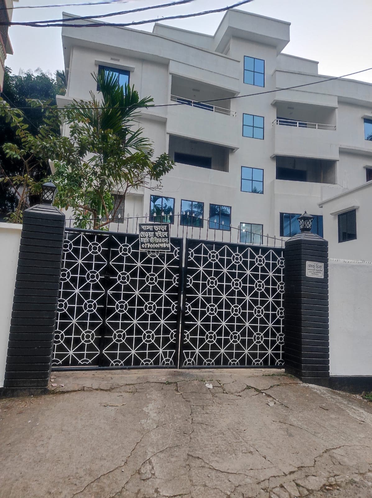 Front gate and multi-storey white building of Arzu Villa Flat 8, daytime, with the black ornamental gate, 'বাসা ভাড়া দেওয়া হবে' rental sign, and palm tree at the entrance