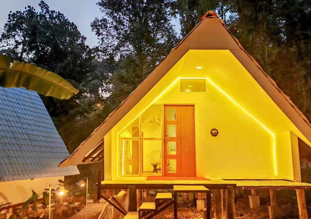 A-frame cottage exterior lit up yellow at dusk, with a wooden entrance door and outdoor seating on the deck