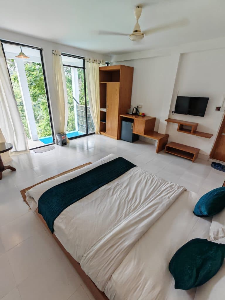 Bedroom with a bed dressed in teal and white linens, wardrobe and TV console, and a glass door opening onto a private plunge pool