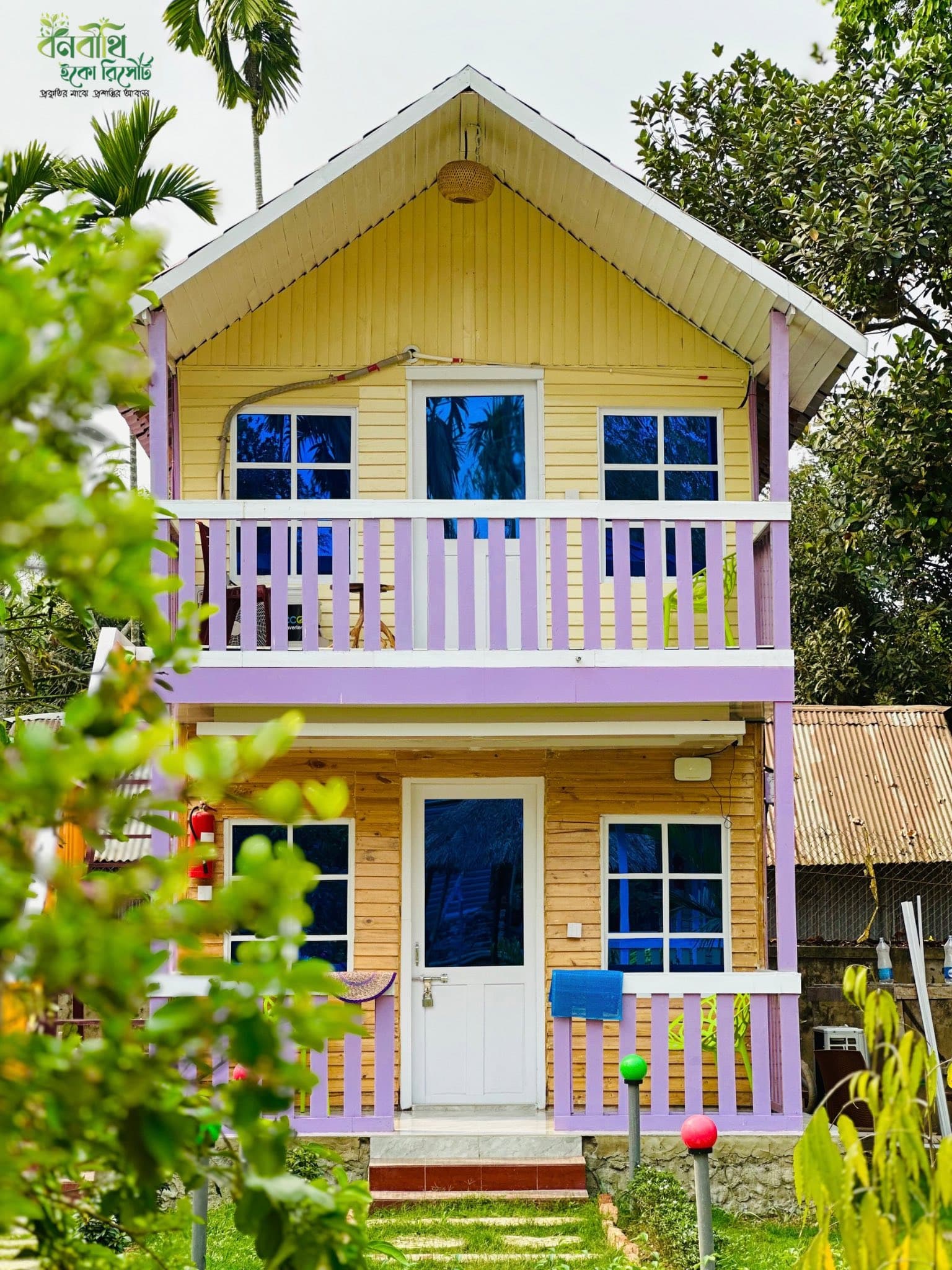 Two-storey duplex cottage building (rooms 103-104) at Banabithi Eco Resort with yellow siding and lavender balcony railings