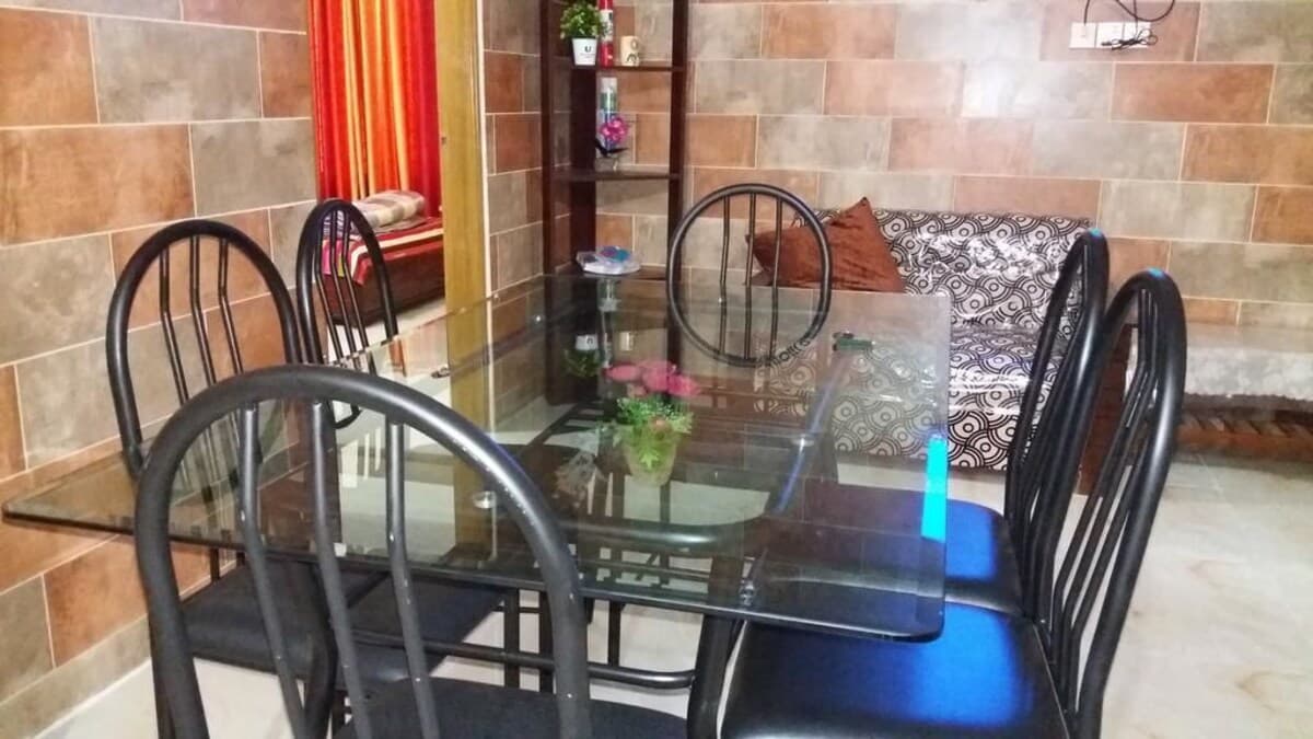 Dining and lounge area with a glass-top table, black metal chairs, a patterned sofa, and a corner display shelf against textured tiled walls