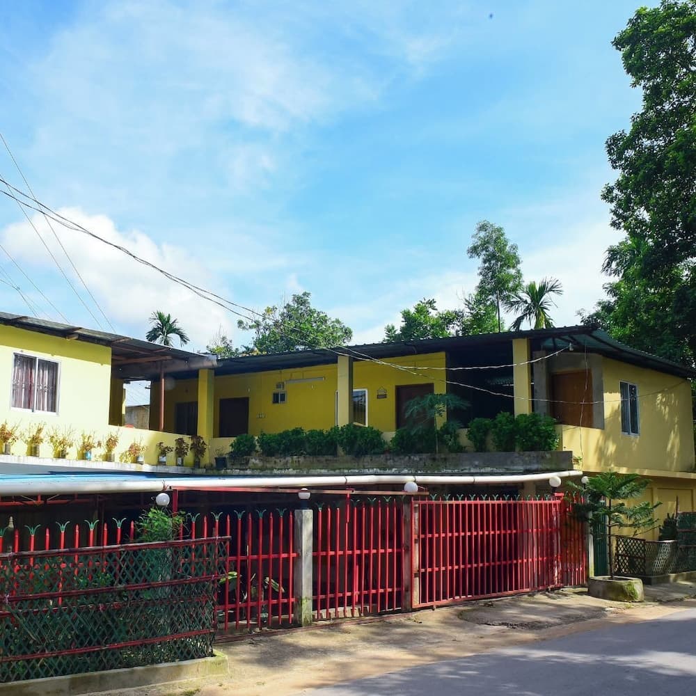 Close-up exterior view of the yellow building's upper-floor balcony and covered porch lined with potted plants, seen over the red perimeter fence