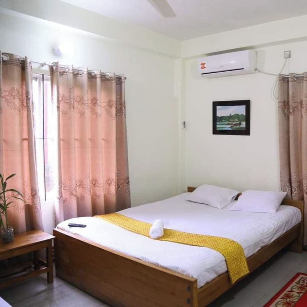 Bedroom with a wooden double bed, dusty-pink patterned curtains, a wall-mounted split AC unit, and a small landscape painting above the headboard