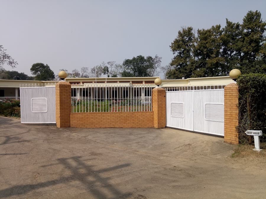 Entrance gate to the guest house compound: two red-brick pillars with pale ball finials flanking white metal gate panels, with a small white arrow signpost reading GUEST HOUSE standing at the roadside on the right, and the long cream single-storey building with a red-framed clerestory window band visible behind the railings