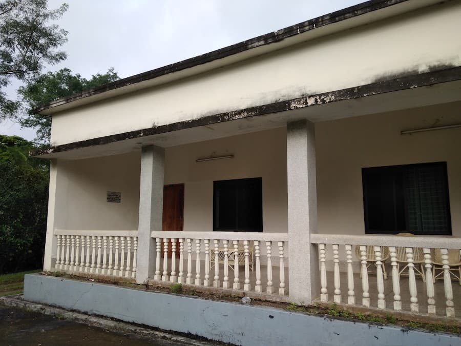 Weathered elevation of the guest house veranda on an overcast day, with a mildew-streaked projecting roof beam, terrazzo-clad square columns, cream turned-baluster railing on a grey-blue painted plinth, a wooden room door with a small Bangla plaque beside it, shuttered windows, and wicker chairs set out at the far end