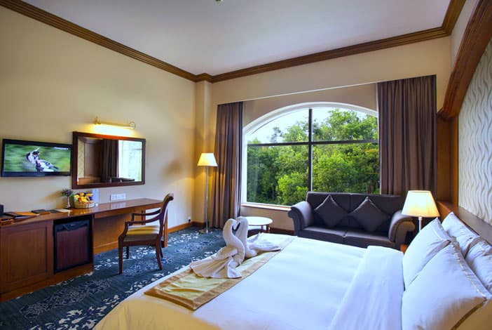 A King Deluxe room at Grand Sultan with a made-up bed, a sofa by an arched window overlooking trees, and a work desk