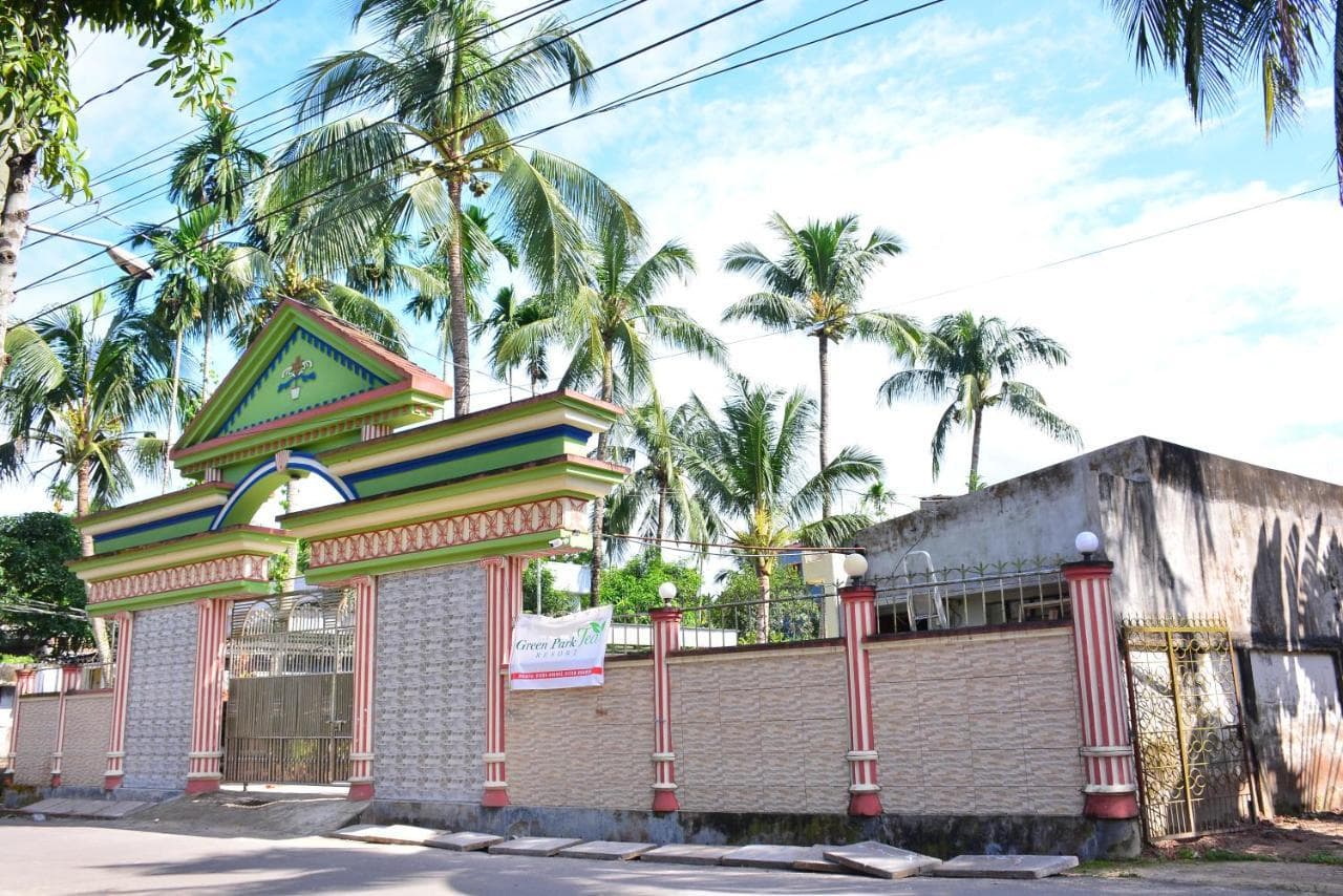 Green-and-red decorative entrance gate with a banner reading "Green Park Tea Resort" hung on the boundary wall, palm trees behind