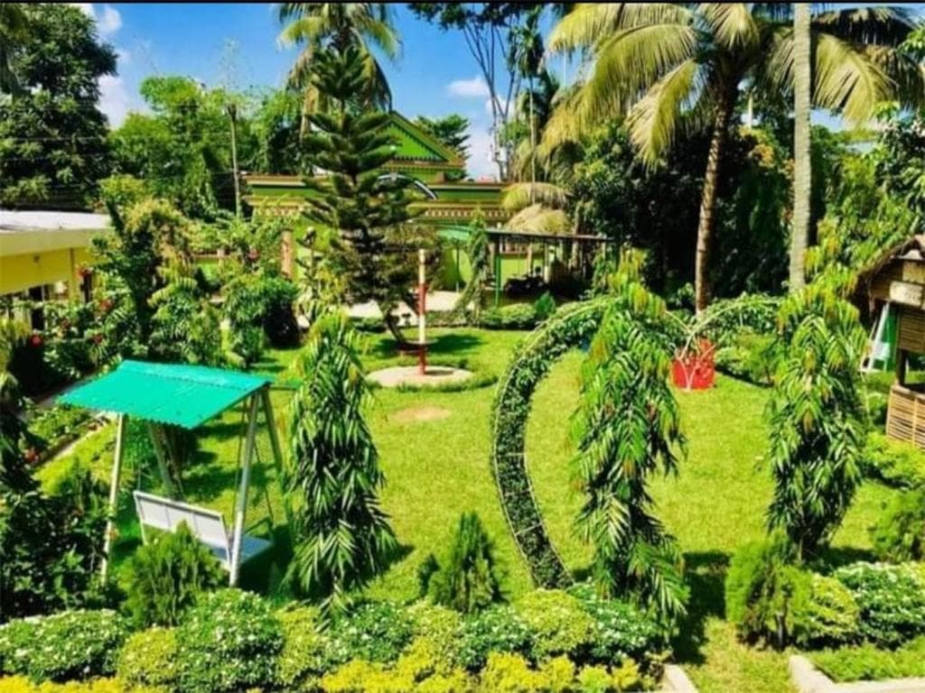 Ground-level view of the resort's garden with a teal-roofed swing bench, ornamental hedges, and the green-trimmed building visible behind the palms