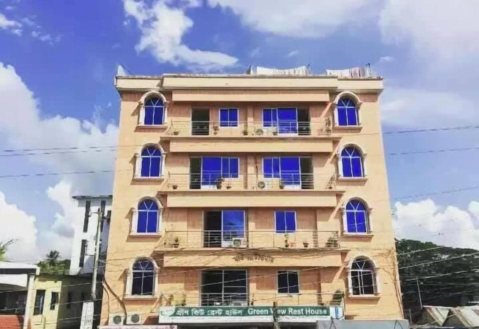 Five-storey cream and beige building with a green signboard across the ground floor reading Green View Rest House in Bangla and English, arched windows with blue-tinted glass, narrow railed balconies on each upper floor and washing hung along the roof parapet