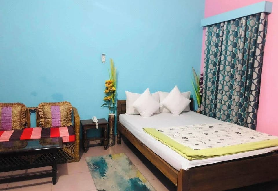 Guest room with a dark wooden double bed and white pillows against a bright teal wall beside a pink painted alcove, a patterned teal curtain, a cane settee with striped cushions, a carved wooden coffee table and a corded telephone on a side table
