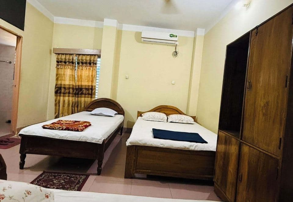Guest room with three wooden-framed single beds on a pink-cream tiled floor, a tall wooden wardrobe, a split air conditioner and small wall fan above, a gold and brown patterned curtain at the window and a maroon floral rug
