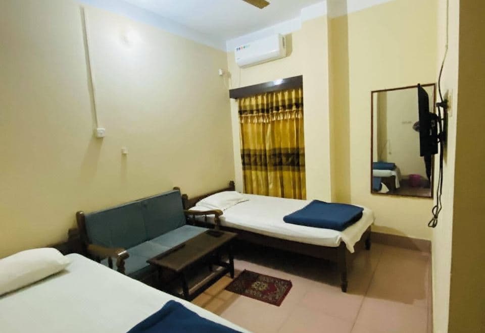 Guest room with a single bed and a wooden-framed settee with teal cushions around a low table, a framed wall mirror and a wall-mounted flat-screen TV on a bracket, a split air conditioner and a gold and brown curtain