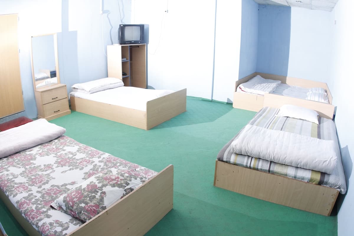 Multi-bed dormitory-style room with pale blue walls, green carpet, four single beds arranged around the room, and a small CRT TV on a corner cabinet