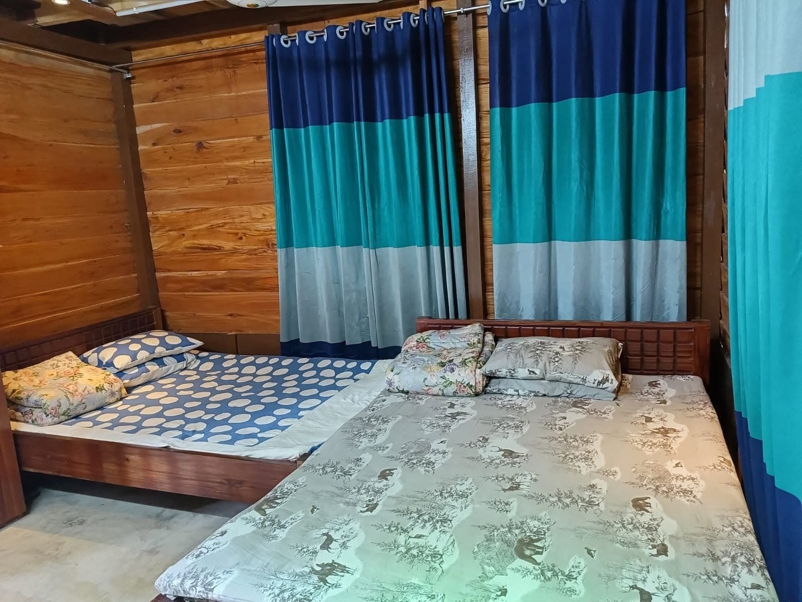 Two wooden-framed beds in a room walled and ceilinged with timber planks, one under a blue-and-white polka-dot spread and one under a beige stag-print spread, with navy, teal and grey colour-blocked curtains behind