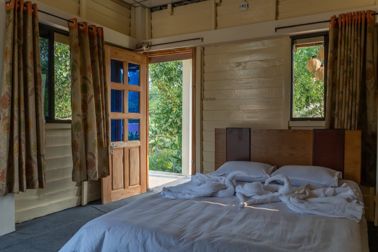 Wood-paneled guest room with floral curtains on two windows, a glass-paned door open to a tea-garden hillside view, and a double bed with white linens and swan-folded towels