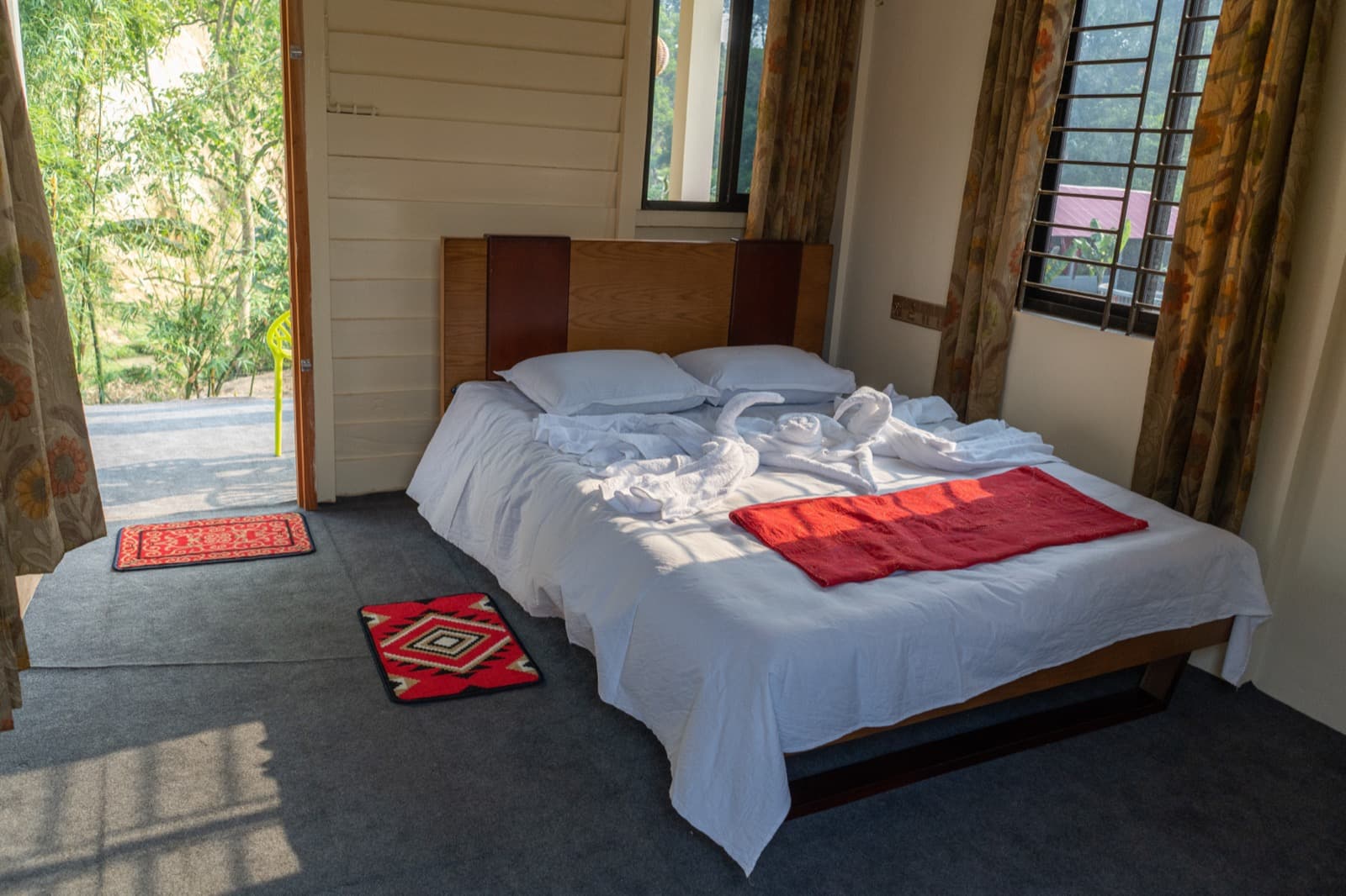 Guest room with a red decorative throw and swan-folded white towels on the bed, patterned rugs on the floor, and an open door leading out to a bamboo grove