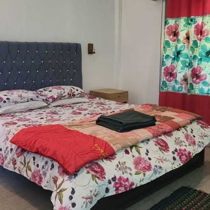 Guest room with a double bed, navy tufted headboard, floral bedspread with a red quilt folded across it, a wooden bedside stool and a floral curtain