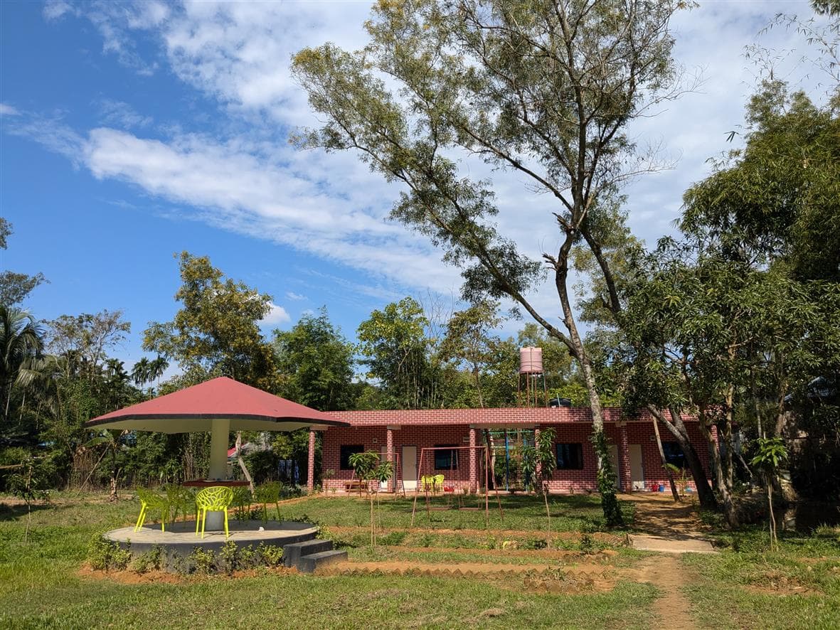 Wide daytime view of the resort's red-brick guest room building with a red umbrella-roof gazebo, yellow chairs, and a children's play area on the lawn in front, water tank on the roof
