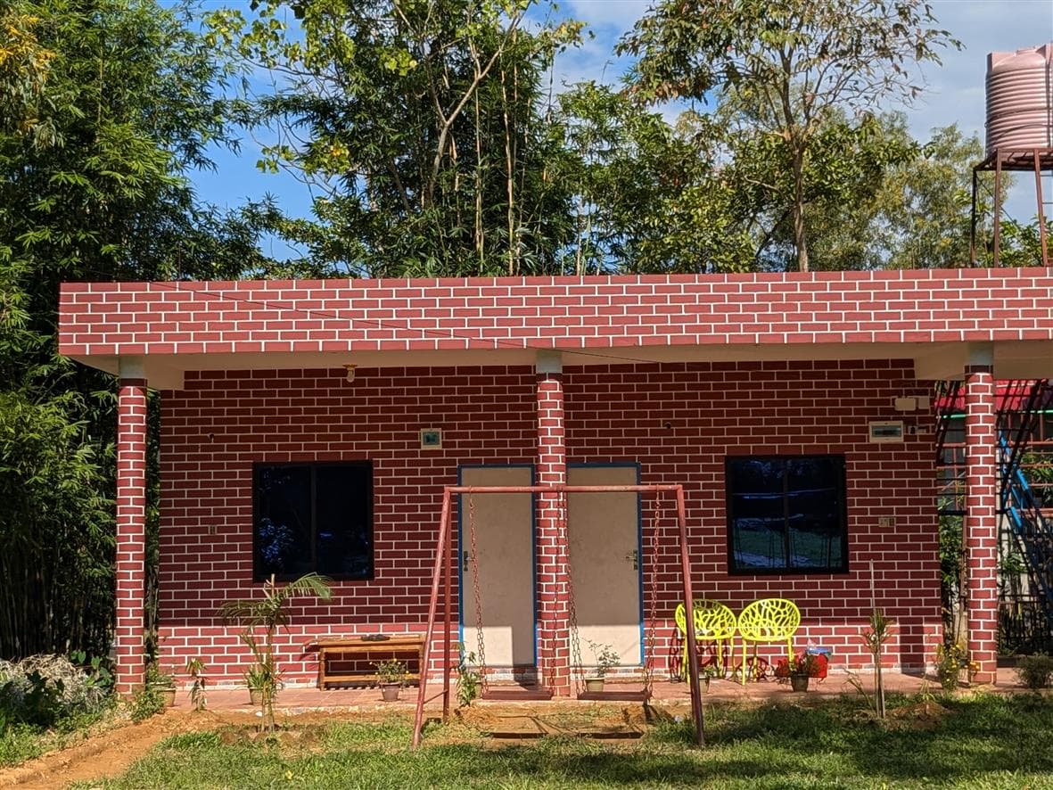 Close-up of the red-brick guest room building facade showing two doors with blue trim, two windows, a swing set, and yellow chairs on the veranda