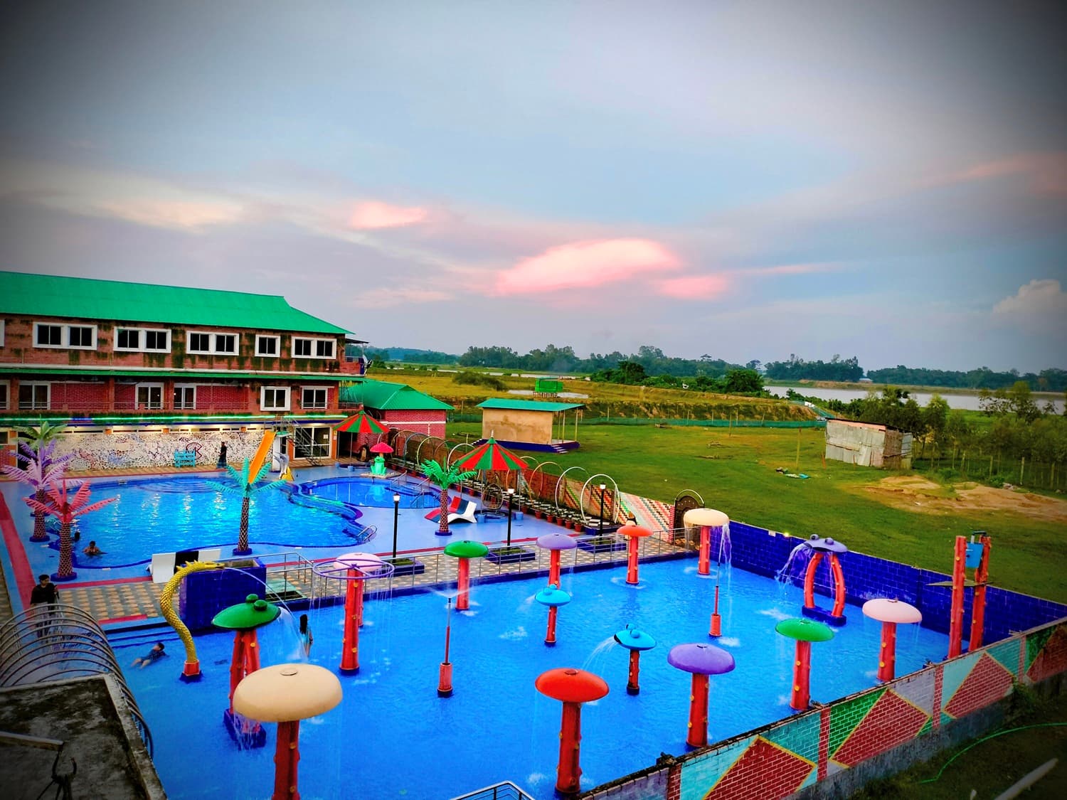 Aerial sunset view of Mukta Nagar Resort's multi-level swimming pool complex with colorful mushroom-shaped water features, the resort's green-roofed brick building, and the adjacent river visible in the background
