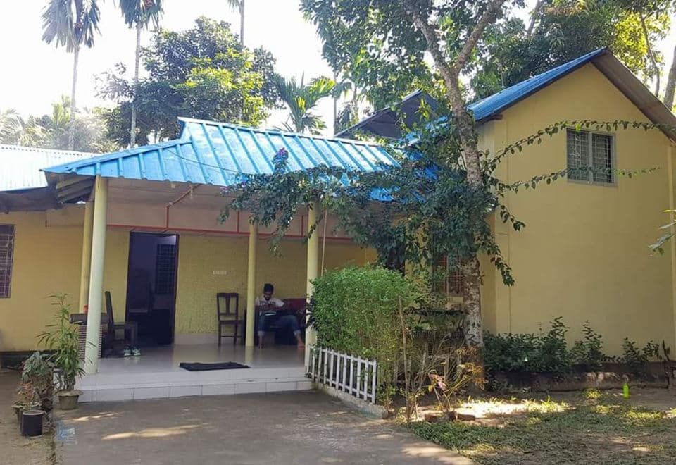 Front of Nilborno Cottage — a single-storey pale-yellow cottage with a bright blue corrugated-metal roof, a deep open veranda on slim columns with a tiled floor and wooden chairs, and a gabled wing at right with a high window, set among betel-nut palms