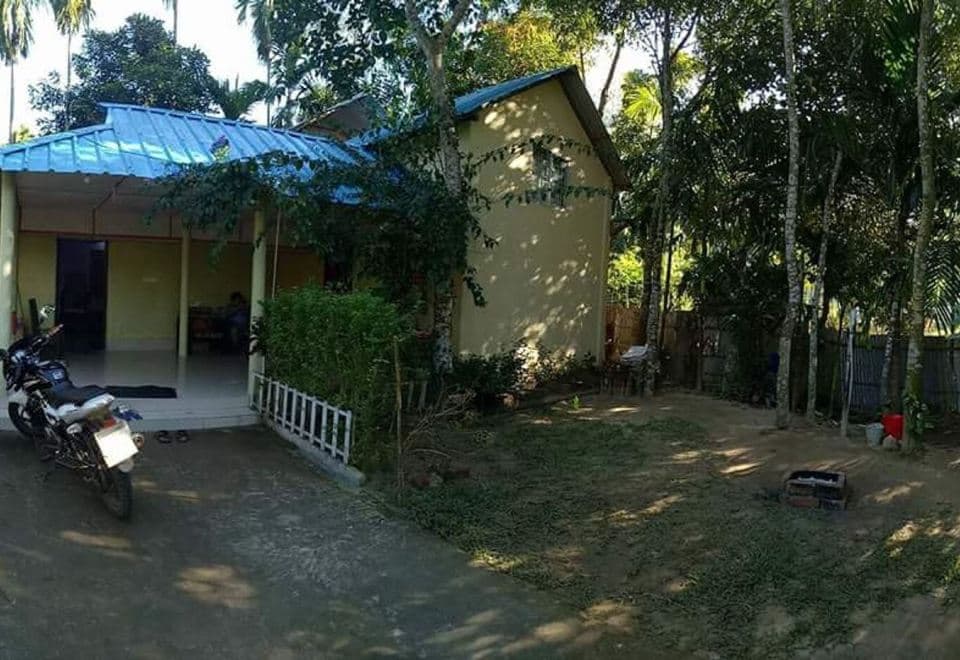 Nilborno Cottage seen from the side across its dirt forecourt, showing the blue corrugated roof over the open veranda, the pale-yellow gable end with a single high window, a motorcycle parked on the drive and sandals left at the veranda step