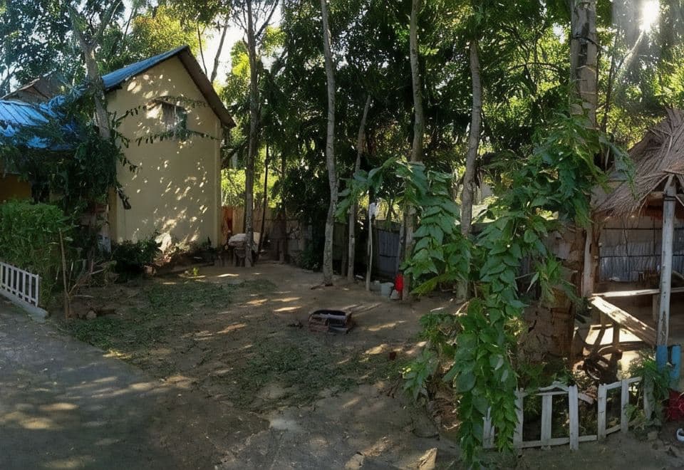 The wooded grounds beside Nilborno Cottage — the cottage's pale-yellow gable end and blue roof at left behind a white picket fence, a shaded bare-earth yard with cut greenery, a bamboo boundary fence, and a thatched-roof open shelter at the right edge