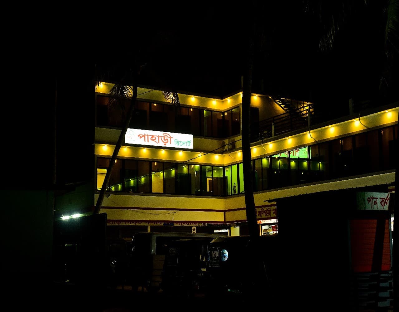 Three-storey Pahari Resort building lit up at night, with the illuminated 'Pahari Resort' signboard on the second-floor facade and vehicles parked in the forecourt