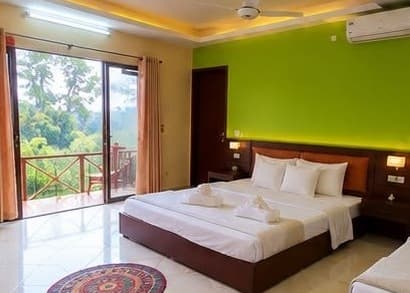 Guest room with a lime-green feature wall, dark-wood double bed made up with white linen and towel swans, ceiling fan and wall-mounted air conditioner, tiled floor, and a sliding glass door opening onto a balcony with a red wooden railing overlooking green hillside trees