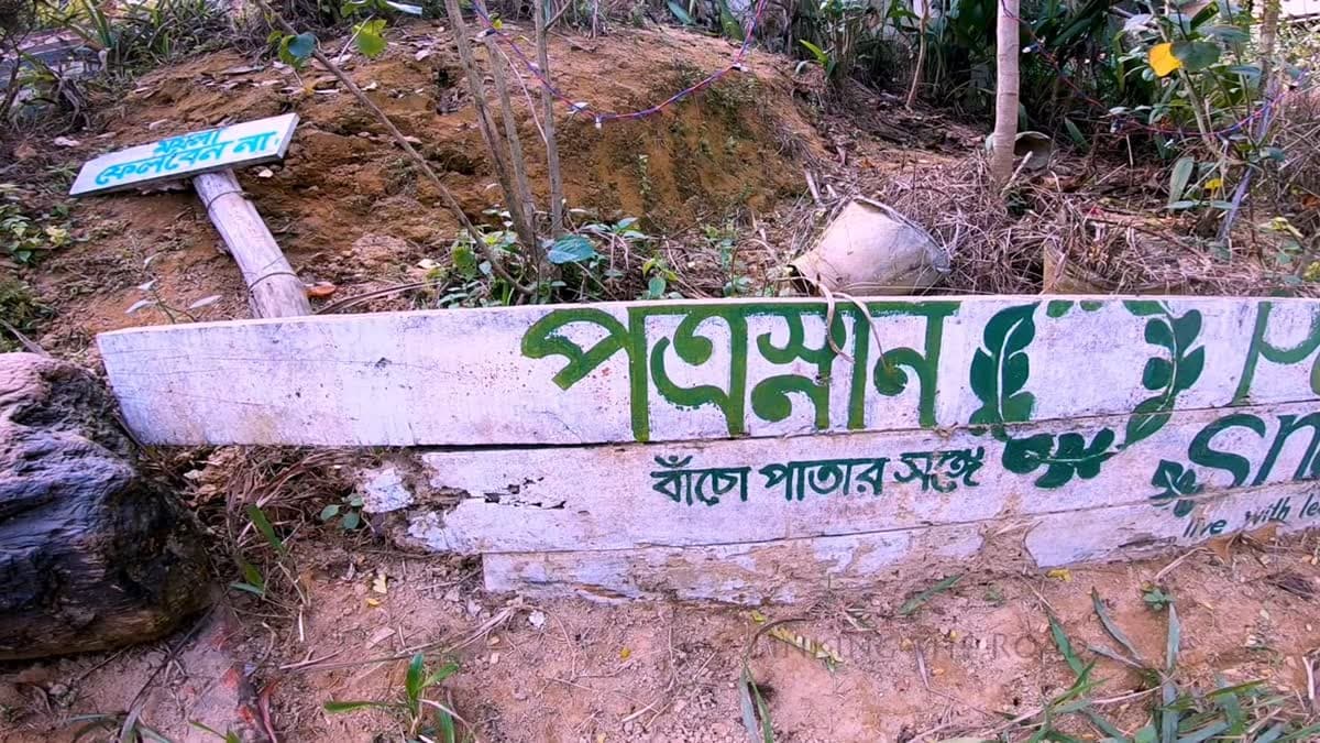 Weathered wooden entrance signboard painted white with green Bangla lettering reading "Patrasnan" and the tagline "live with leaves", set into a dirt embankment