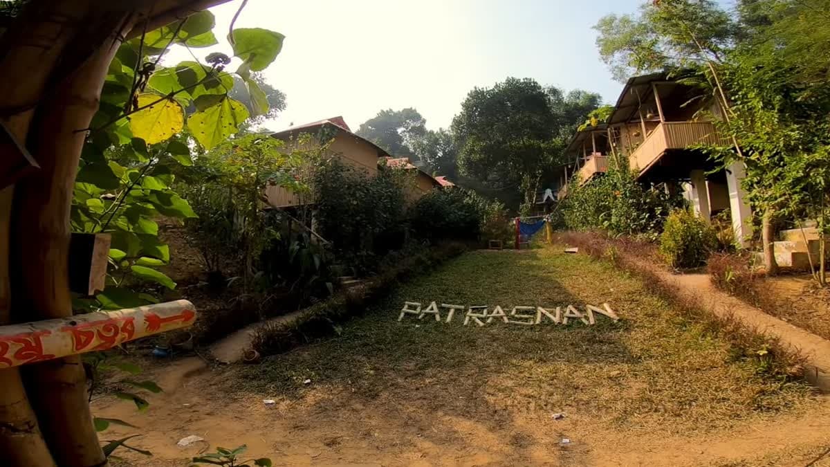 Grass walkway spelling out "PATRASNAN" in white lettering, flanked by two-storey bamboo cottages on stilts, with a hand-painted directional signpost in the foreground