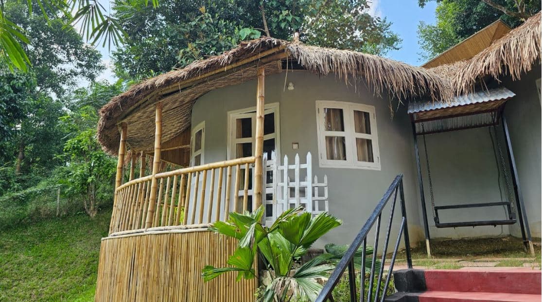 Close-up of a bamboo-and-thatch cottage exterior (Kureghor cottage) with wooden shutters and brick steps