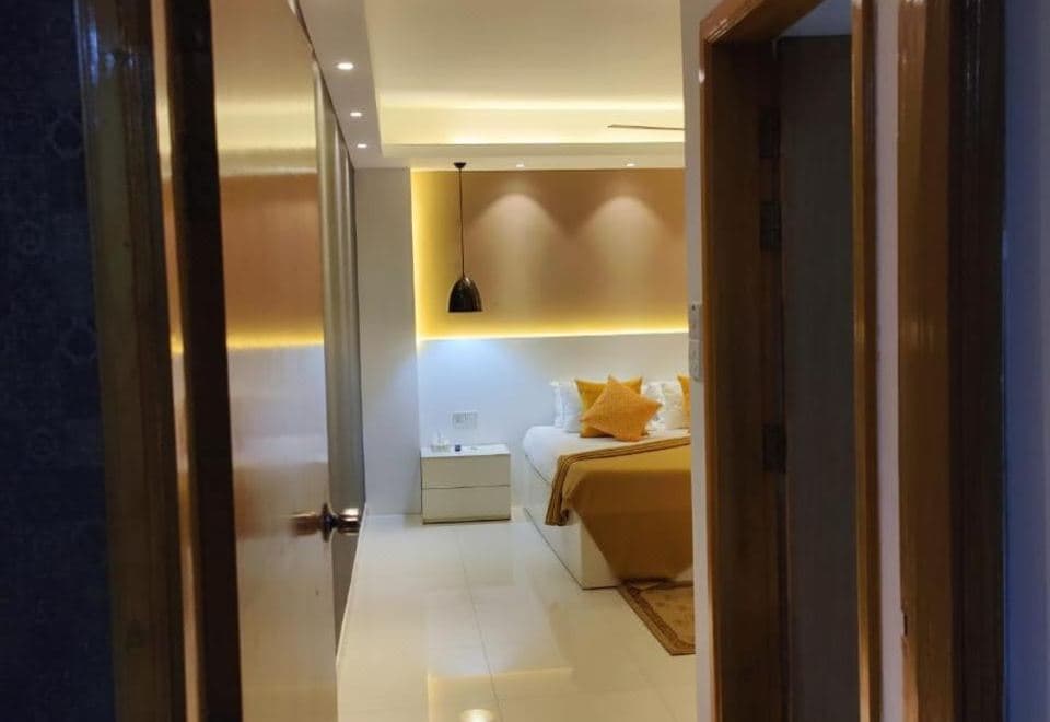 Guest room with a mustard-yellow bedspread, pendant lamp over the headboard, and warm cove lighting seen through the open doorway