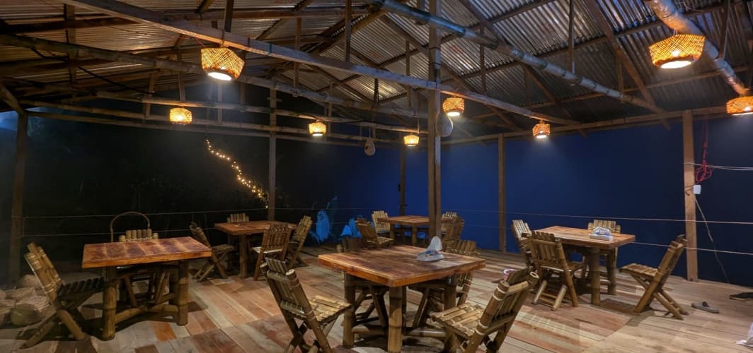 Open-air bamboo-and-tin restaurant pavilion at night, with wooden tables, bamboo chairs, and hanging lantern-style lights