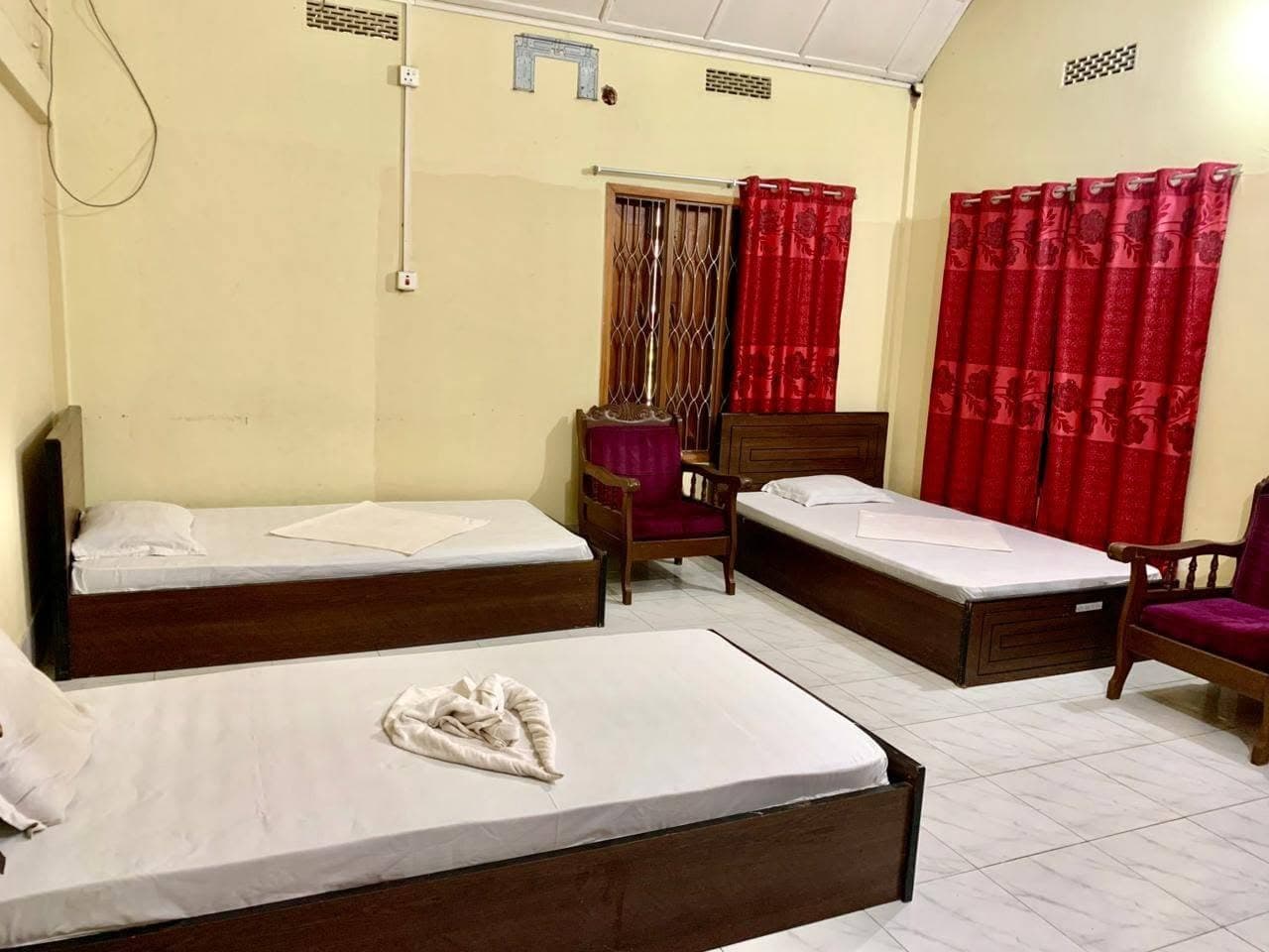 Family room with dark wood-framed single beds made up in white linen, red rose-patterned curtains, two purple-upholstered wooden armchairs and a white tiled floor