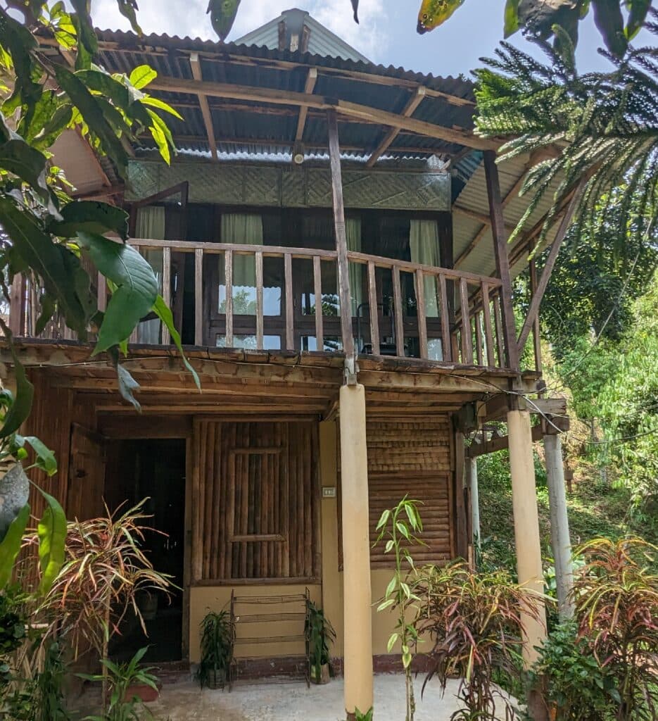A two-storey wooden cottage at Shanti Bari with an upstairs balcony, surrounded by mango trees