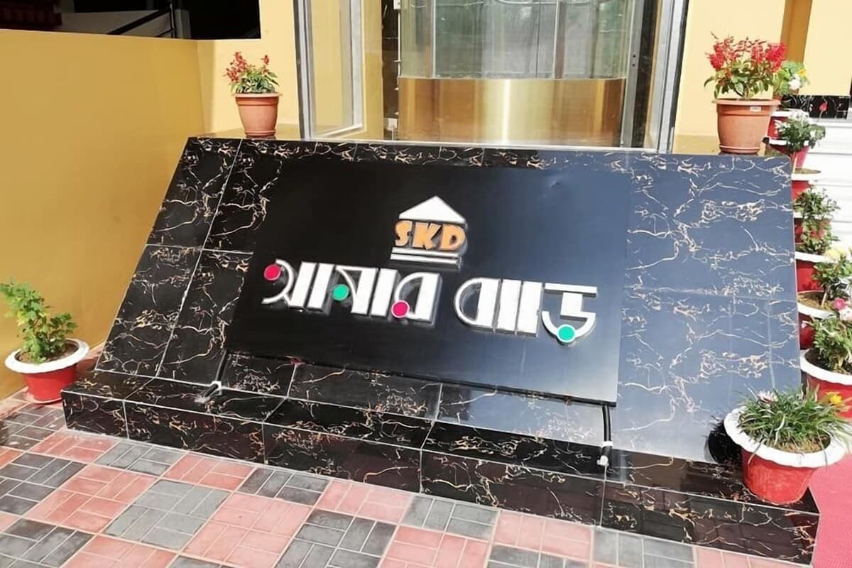 Close-up of the black marble entrance monument engraved with the SKD logo and "আমার বাড়ি" (Amar Bari) lettering, flanked by potted plants at the building entrance