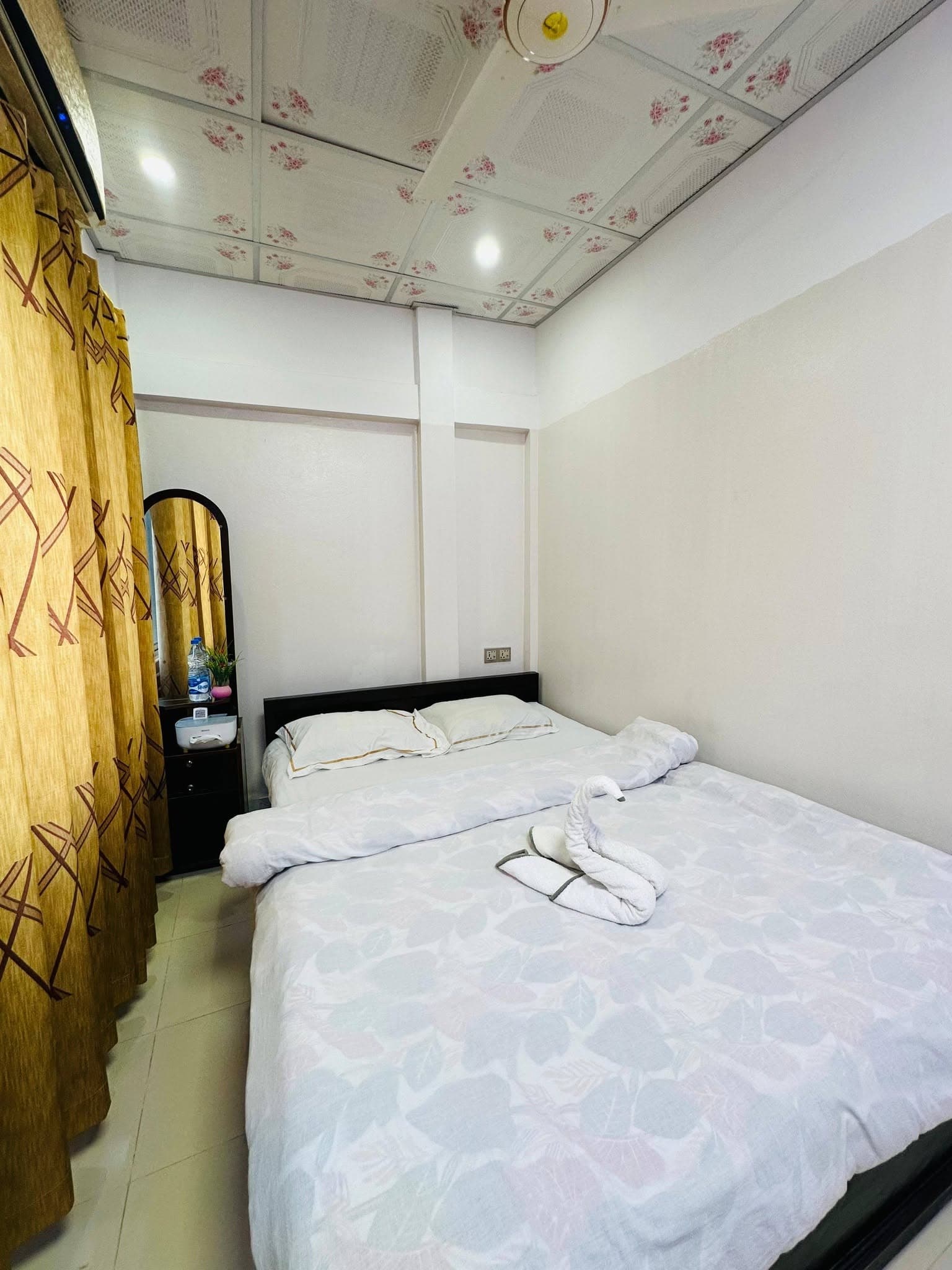 Guest room with a double bed, a towel folded into a swan shape, patterned floral ceiling tiles, and a gold curtain