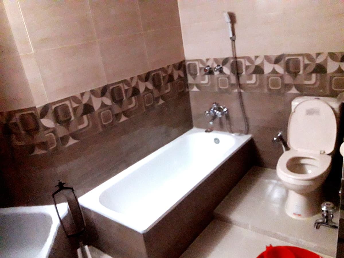 En-suite bathroom at Sreemangal Cottage with a built-in white tub, wall-mounted handheld shower, a Western-style toilet with bidet spray and a washbasin at the left, on beige tile with a brown geometric border course