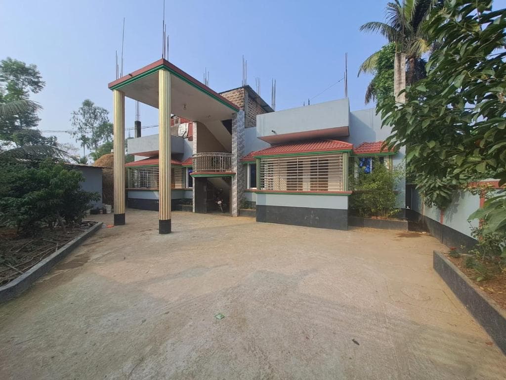 Two-storey concrete guest house with a tall columned carport, red-tiled window awnings with green trim, and exposed roof rebar, fronted by a wide bare-earth driveway with palms and leafy trees on both sides