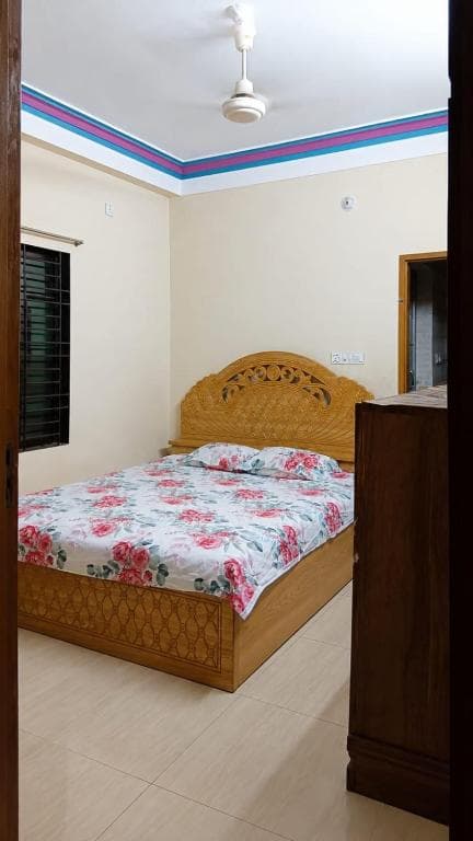 Bedroom seen through an open doorway: a double bed with pink-and-white floral bedding and a carved wooden headboard, a barred window with dark curtain, a ceiling fan, and a pink-and-blue painted border running around the ceiling edge