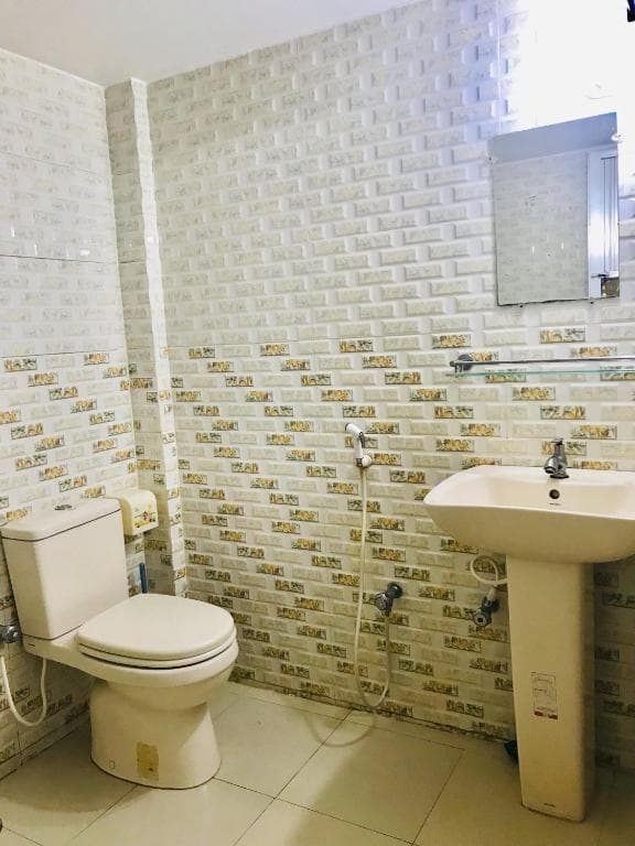 Bathroom tiled floor-to-ceiling in cream brick-pattern tile, with a Western-style toilet, a white pedestal basin with chrome tap, a wall mirror with glass shelf, and a hand-held bidet shower hose