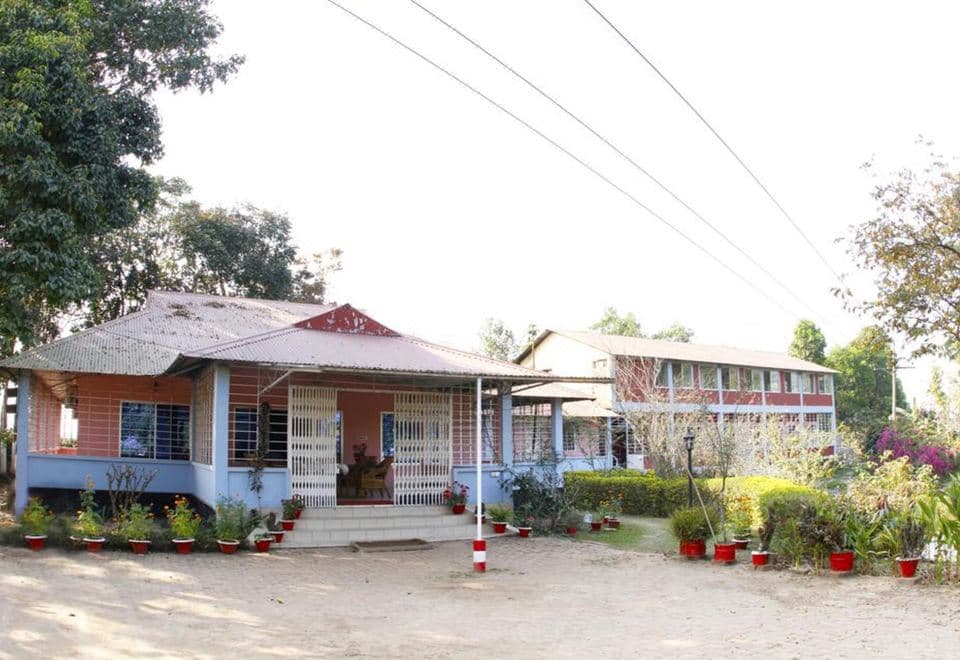 Front entrance of Sreemangal Resort — single-story pink-and-blue guest wing with a red tin roof and grille-covered veranda, connected to the two-story red brick block, with a paved courtyard and potted plants lining the entrance path