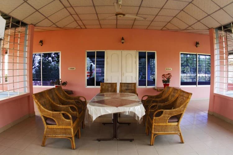 Covered salmon-pink veranda at Sreemangal Resort furnished with a large round dining table and six woven cane armchairs, French doors and grilled windows along the back wall