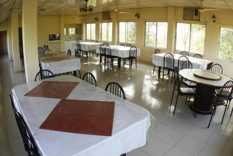 Rooftop dining hall at Sreemangal Resort with rows of white-clothed tables and black chairs under ceiling fans, floor-to-ceiling windows overlooking the surrounding trees