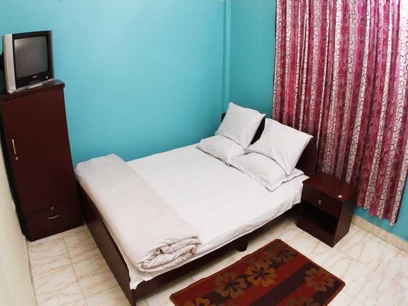 Guest bedroom at Sreemangal Resort with turquoise walls, a double bed with white bedding, a dark wood wardrobe and nightstand, a CRT television, and floral pink curtains