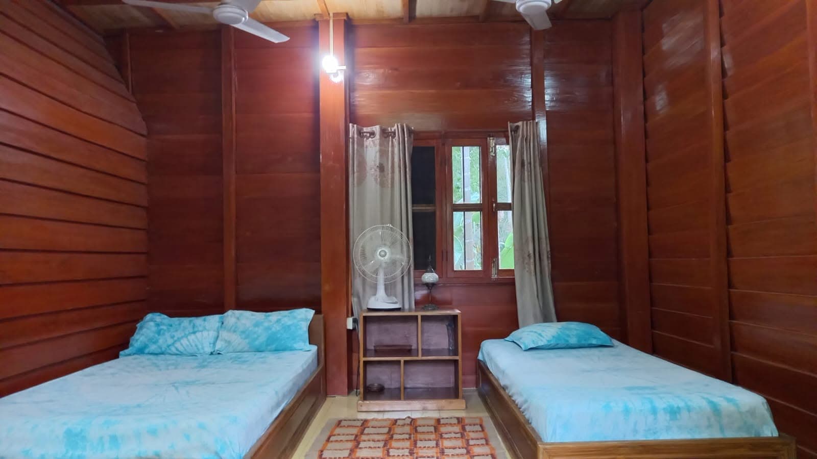 Guest room lined floor to ceiling in varnished wood, with a double and a single bed under pale blue covers, two ceiling fans, a pedestal fan on a low shelf unit, and a shuttered window looking onto green foliage