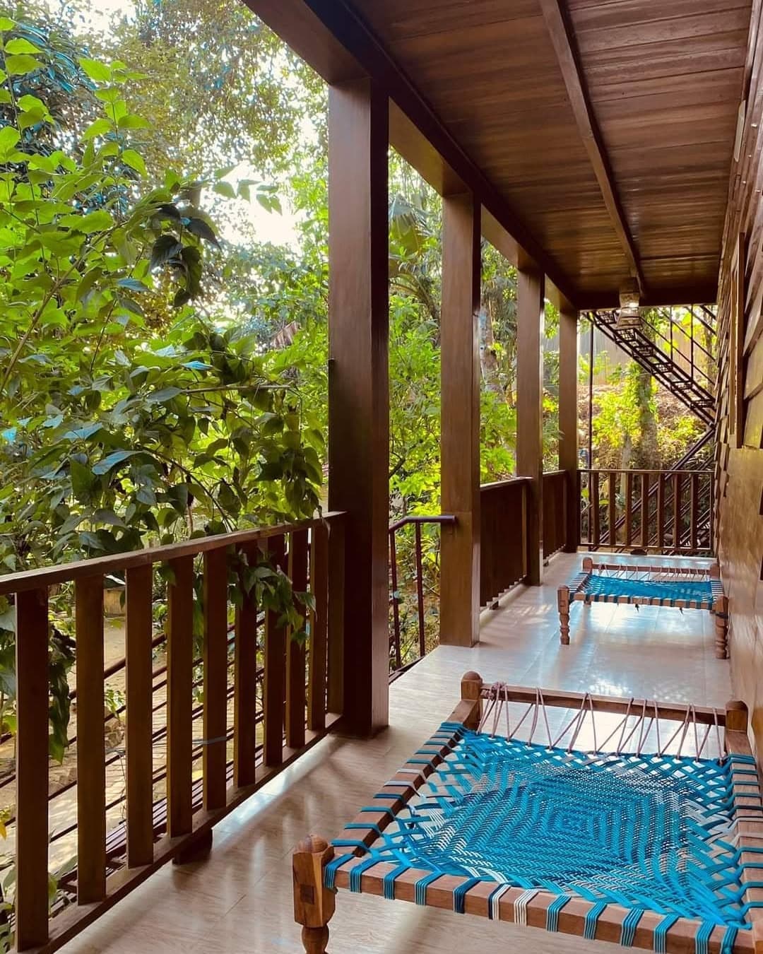 Covered upper veranda with a varnished timber ceiling and baluster railing, two traditional woven charpai daybeds strung in bright blue standing on a tiled floor, and a steel staircase at the far end