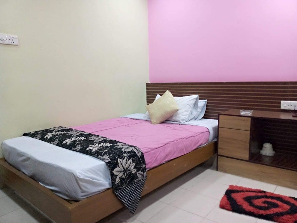 A double guest room at Sultan Restaurant & Guest House with a pink bedspread and pink accent wall, a horizontal wood-slat headboard, a bedside cabinet and a red patterned rug on white tiled floor